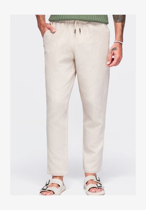 Mand iført off-white teksturerede bukser med snøre i taljen, grøn ribstrikket sweater, hvide sandaler og tatoveringer på synlige hænder og underarme.