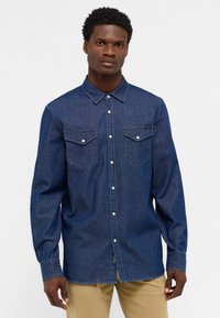 Granatowa koszula dżinsowa z długim rękawem, z kołnierzykiem zapinanym na guziki, dwiema kieszeniami na piersi z klapami oraz zaokrąglonym dołem. Srebrne guziki zdobią przód.