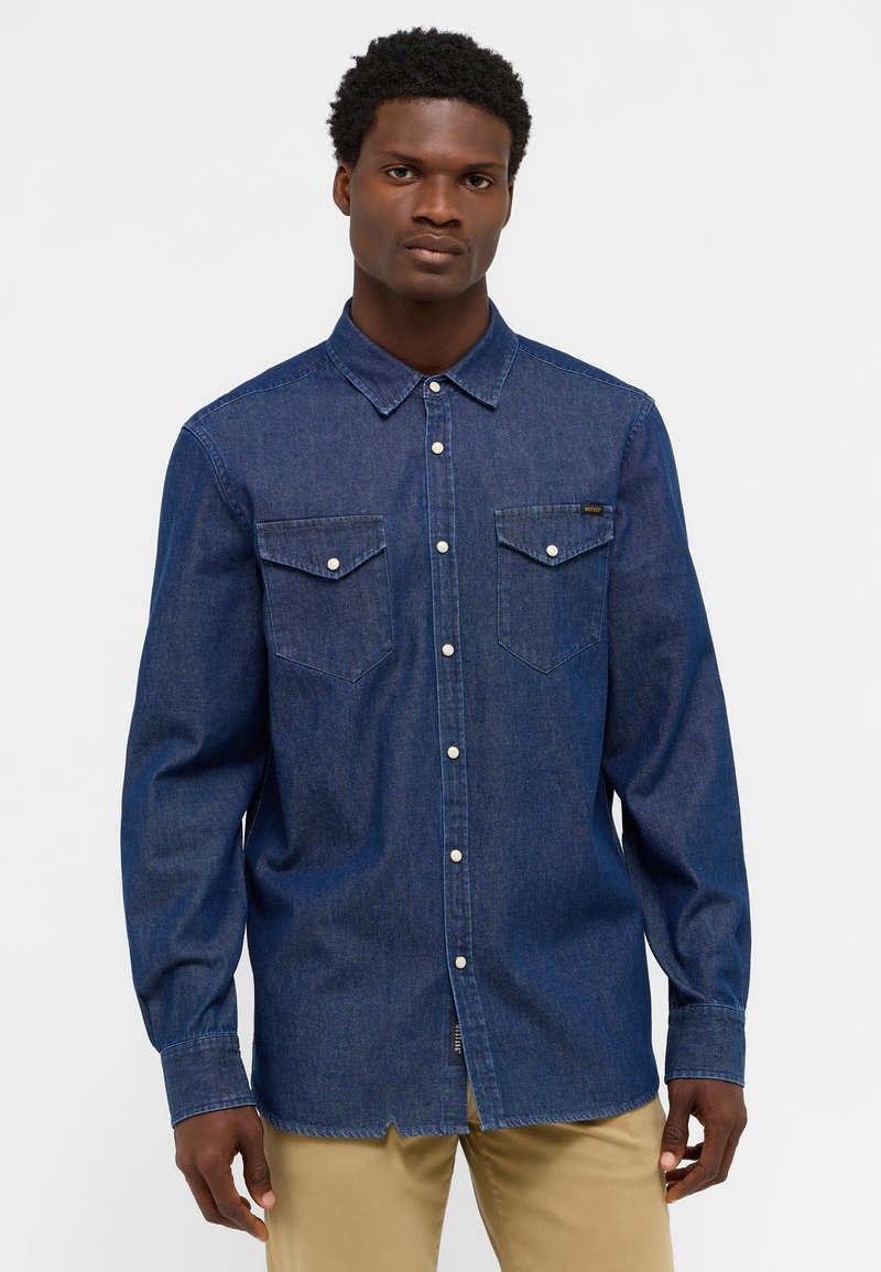Granatowa koszula dżinsowa z długim rękawem, z kołnierzykiem zapinanym na guziki, dwiema kieszeniami na piersi z klapami oraz zaokrąglonym dołem. Srebrne guziki zdobią przód.