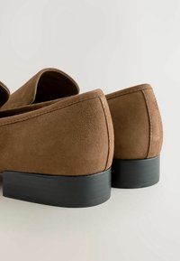 Paire de mocassins en daim marron avec de petits talons noirs, présentés de dos et de côté sur un fond clair uni.