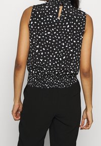 Svart ärmlös topp med vitt prickmönster, samlad halsringning och elastisk midja. Modellen bär matchande svarta byxor.