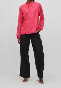 Blusa de patrón de chevron rosa con un cuello alto y con volantes, y mangas largas, combinada con pantalones negros holgados y sandalias de tacón transparente.