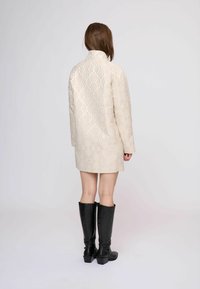Beige gestippelde jas met geometrisch ontwerp, met een hoge kraag en lange mouwen, gecombineerd met zwarte kniehoogte laarzen.