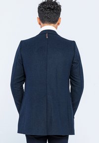 Marinblå skräddarsydd kavaj med en enda vent, strukturerade axlar och synliga sömdetaljer. Har en kontrasterande vit kragefoder.