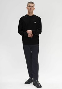 Schwarzer Pullover mit langen Ärmeln, Rundhalsausschnitt und kleinem goldenen Logo auf der Brust, kombiniert mit dunklen Hosen und schwarzen Schuhen. Einfaches Design.