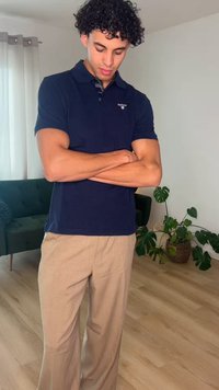 Młody mężczyzna o kręconych włosach, ubrany w granatową koszulkę polo i beżowe spodnie, stoi w pomieszczeniu z założonymi rękami, obok zielonej sofy i doniczkowych roślin.