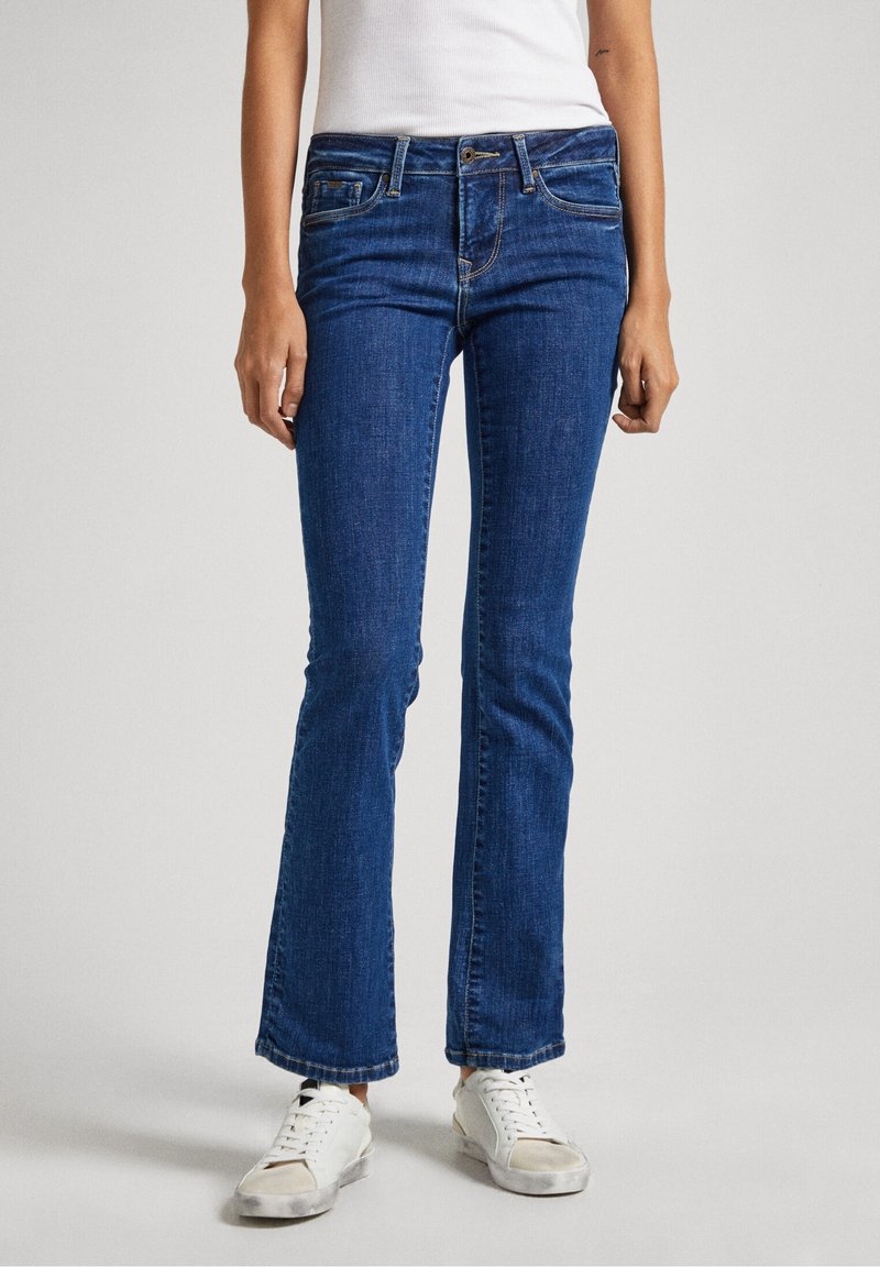 Jean évasé en denim bleu léger avec une coupe régulière, cinq poches et des surpiqûres contrastantes discrètes, associé à des baskets blanches.