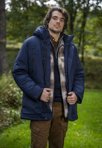 Marineblaue Steppjacke mit Kapuze, ausgestattet mit Reißverschluss und Knöpfen. Trägt darunter ein karierte Flanellhemd und einen dunklen Pullover. Braune Cargohose.