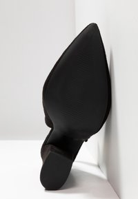 Chaussure à talon haut en daim noir avec un bout pointu et une semelle texturée. Le talon est fin et la chaussure présente une finition lisse et mate.