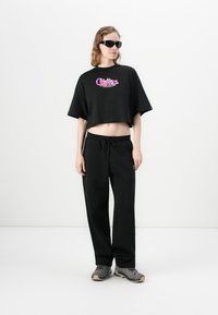 Kvinde i sort cropped grafisk t-shirt, løse sorte bukser, grå sneakers og sorte solbriller, der står mod en hvid baggrund.