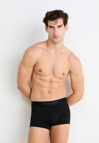 Boxer-briefs noirs avec une ceinture élastique Calvin Klein, présentant une texture lisse et une coupe ajustée. Portés par un modèle sur un fond neutre.