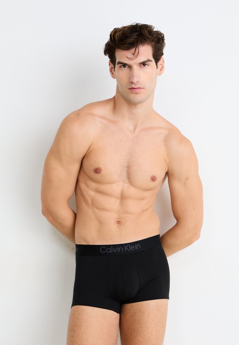 Boxer-briefs noirs avec une ceinture élastique Calvin Klein, présentant une texture lisse et une coupe ajustée. Portés par un modèle sur un fond neutre.