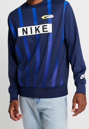 Osoba ubrana w granatową bluzę z pionowymi niebieskimi paskami i białymi logo Nike na piersi i rękawie oraz w jasnoniebieskie dżinsy.