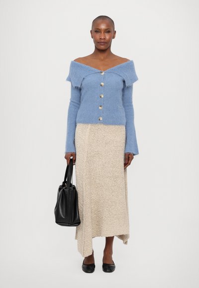 Light blue off-shoulder cardigan with large buttons, paired with a beige textured midi skirt. Black handbag and flat shoes complete the look.