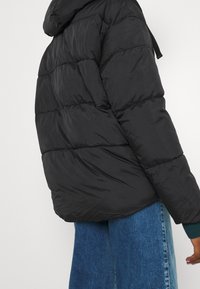 Giacca a vento nera con cappuccio, caratterizzata da un design trapuntato, polsini elastici e una texture liscia e lucida. Indossata sopra a dei jeans blu.