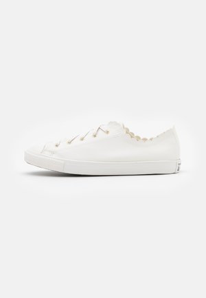 White leather sneaker with scalloped edges, flat lace-up design, gold eyelets, textured toe cap, and white rubber sole.