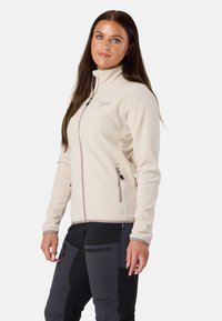 Beige fleece-takki korkealla kauluksella, vetoketjulliset taskut ja kontrastivärinen pylväs. Yhdistetty mustiin housuihin, joissa on vetoketjuelementtejä.