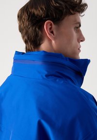 Giacca impermeabile blu con colletto alto, texture liscia e dettagli delle cuciture. Presenta una zip sul colletto per una vestibilità regolabile.