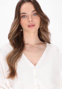 Witte v-hals top met een zachte textuur, met een knoopsluiting aan de voorkant. Lange, golvende haren en ronde pareloorbellen maken de look compleet.