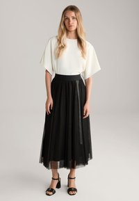 Haut blanc fluide avec de larges manches associé à une jupe en tulle noire à plusieurs niveaux. Sandales à talons blocs noires avec des brides à la cheville.