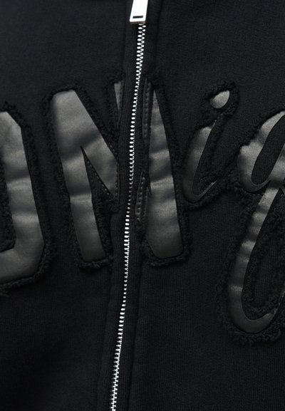 Κοντινή λήψη μαύρου υφάσματος με ασημένιο φερμουάρ και μεγάλους υφασμάτινους μαύρους χαρακτήρες που είναι ραμμένοι γύρω από το φερμουάρ σε ένα ένδυμα.