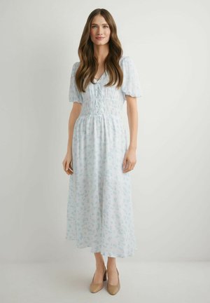 Kobieta w jasnoniebieskiej kwiecistej sukience midi z bufkami na rękawach i beżowych szpilkach, stojąca na tle jednolitego jasnego tła.