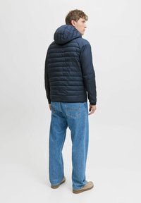 Marineblå polstret jakke med hætte, der har vandrette syninger og lange ærmer, parret med løstsiddende blå denimjeans og beige sko.