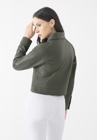 Giacca in pelle verde oliva con un design cropped, colletto a scatto e dettagli di cucitura visibili, indossata su pantaloni bianchi attillati.