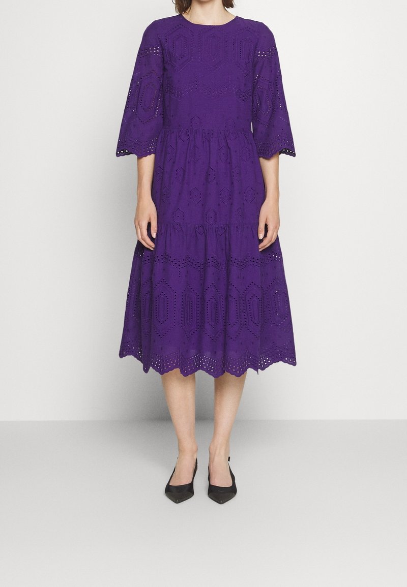 Femme portant une robe violette jusqu'aux genoux avec des motifs à œillets et des manches trois-quarts, associée à des escarpins noirs à bouts pointus.