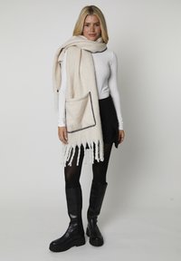 Écharpe beige clair avec une texture en chevron, dotée d'une poche avant bordée de broderie noire et d'extrémités à franges.