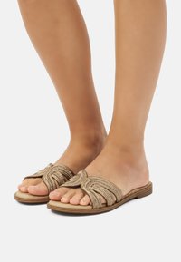 Sandalias planas doradas con un diseño de tira ondulada; adornadas con acentos de cuentas. Material suave, punta abierta y una suela de madera baja.