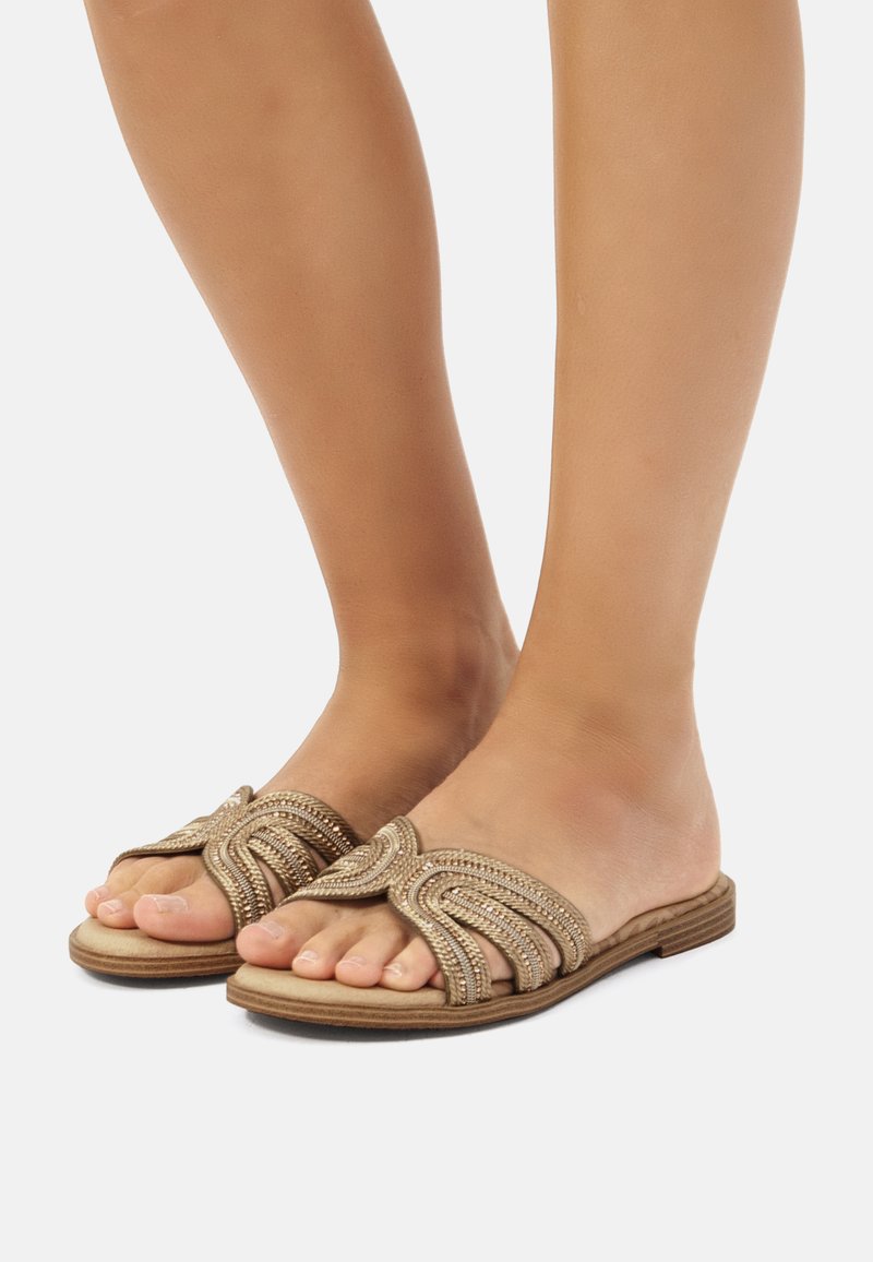 Sandalias planas doradas con un diseño de tira ondulada; adornadas con acentos de cuentas. Material suave, punta abierta y una suela de madera baja.
