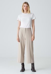 Pantalon large beige avec des plis à l'avant, associé à un t-shirt blanc ajusté. Le mannequin porte des bottines châtaigne. Fond neutre.