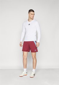 Witte lange mouwen shirt met zwart Adidas-logo; bordeauxrode shorts met blauwe accenten; witte sportschoenen. Schoon ontwerp, getailleerde stijl.