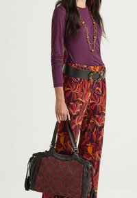 Lila Langarmshirt kombiniert mit einer Hose im Blumenmuster in Rot, Orange und Lila. Abgerundet wird das Outfit durch einen breiten schwarzen Gürtel und eine schwarze Handtasche mit einer strukturierten Oberfläche.