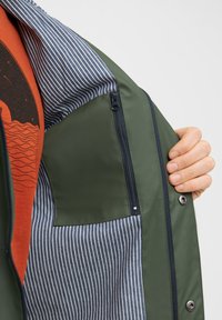 Giacca verde con una fodera a righe blu e bianche, tasca con zip e bottoni a pressione, realizzata in materiale esterno liscio e resistente all'acqua.