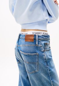 Osoba nosi modré džínsy s koženou štítkom „Tommy Jeans“ a bielym pásom viditeľným nad džínsami, na bielom pozadí.