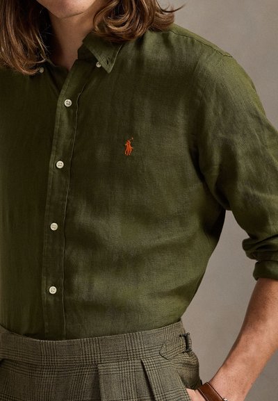 Olivovo zelená ľanová košeľa na zapínanie na gombíky s bielymi gombíkmi a malým oranžovým emblémom polotrička; v kombinácii so sivými kockovanými nohavicami s pevne tvarovaným pásom.