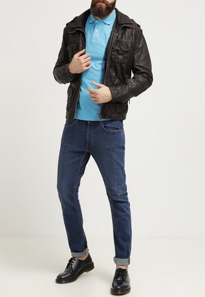 Homme portant une veste en cuir noire, un polo bleu, un jean bleu foncé retroussé et des chaussures noires à lacets, debout devant un fond blanc.