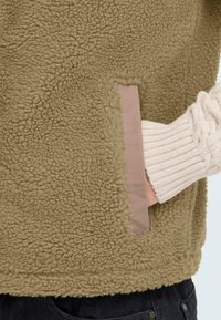 Fleece jacket in olive green with a textured, fluffy surface. Features a side pocket lined with a soft pink accent. Cable-knit sleeve detail visible.