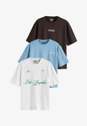 Trois t-shirts à manches courtes pliés en marron, bleu clair et blanc, chacun avec un texte à thème urbain imprimé sur le devant.
