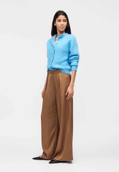 Γυναίκα με ανοιχτό μπλε χνουδωτό ζακέτα, καφέ παντελόνι με φαρδιά μπατζάκια και μαύρα μυτερά παπούτσια, όρθια και κοιτάζοντας προς τα πλάγια σε λευκό φόντο.