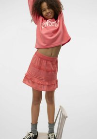 Koralfarvet sweatshirt med fed hvid tekst, parret med en koralfarvet lagdelt flæseskirt. Grå sokker med bølgede kanter og sorte sneakers.