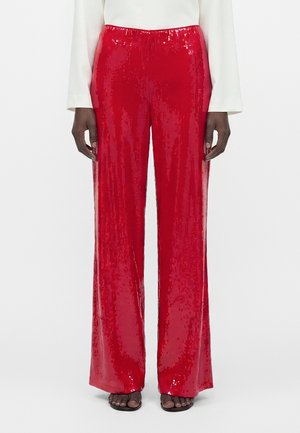Femme portant un pantalon rouge à sequins, taille haute et jambes larges, associé à un haut blanc à manches longues et des sandales noires ouvertes à l'avant.