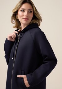 Giacca con cappuccio di colore navy con zip frontale, maniche a raglan, tasche laterali e dettagli in metallo argentato. Tessuto liscio, design casual.