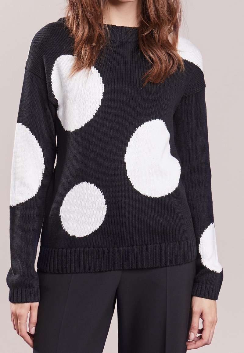 Pull en maille noire avec de grands pois blancs irréguliers porté par une personne aux cheveux châtain mi-longs, assorti à un pantalon noir.