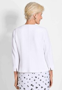 Weißer strukturierter Blazer mit Dreiviertelärmeln, geradem Saum und seitlichen Schlitzen, kombiniert mit einem gemusterten Rock mit schwarzen Designs.