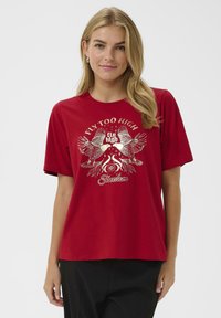 Raudona medvilninė marškinė su trumpomis rankovėmis, turinti metalinę grafikos dizainą su dviem paukščiais ir tekstu „Skristi per aukštai“ meninėmis raidėmis.