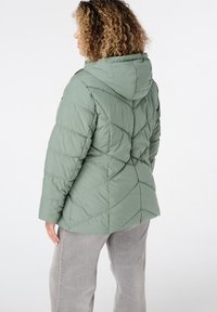 Puffy groene jas met capuchon, met diagonale gewatteerde patronen en elastische manchetten. Draag over lichtgrijze broek.