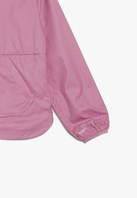 Veste coupe-vent rose clair avec un col rond, des poignets élastiques et une poche avant. Fabriquée en matériau doux et léger.